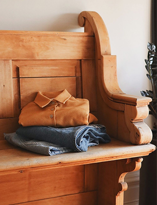 A light brown hall bench with 3 pieces of cloths on it.